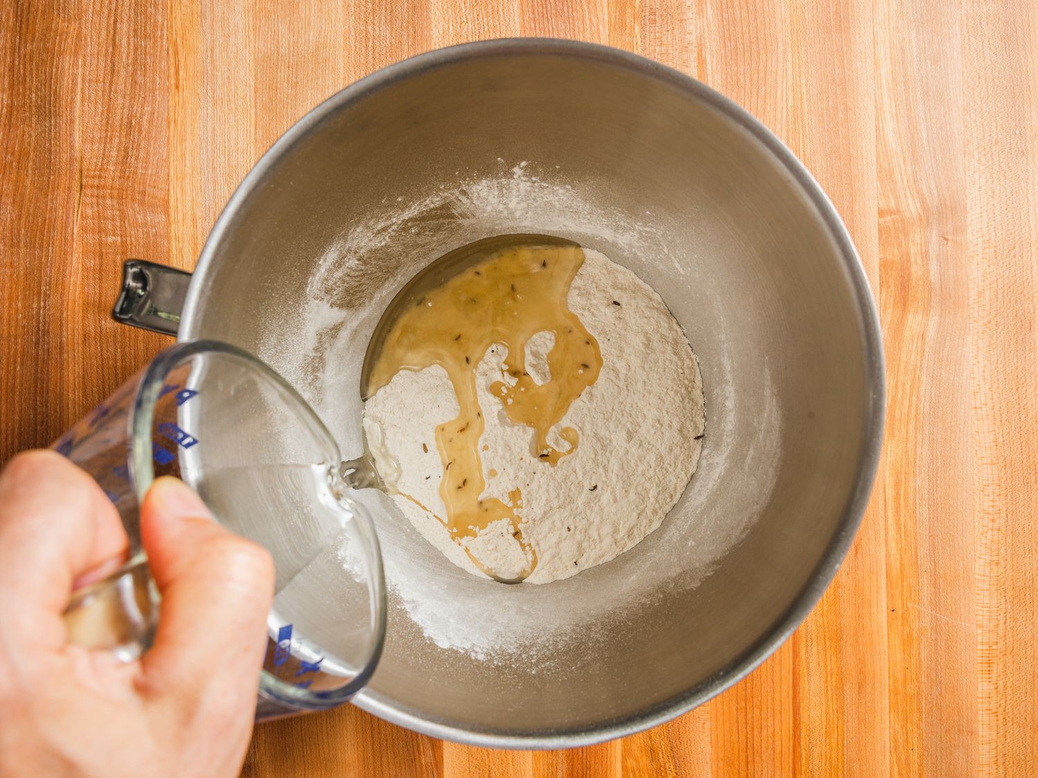 Overhead view of dry mix for samosa dough in a stand mixer bowl with oil being poured from a measuring cup into the bowl