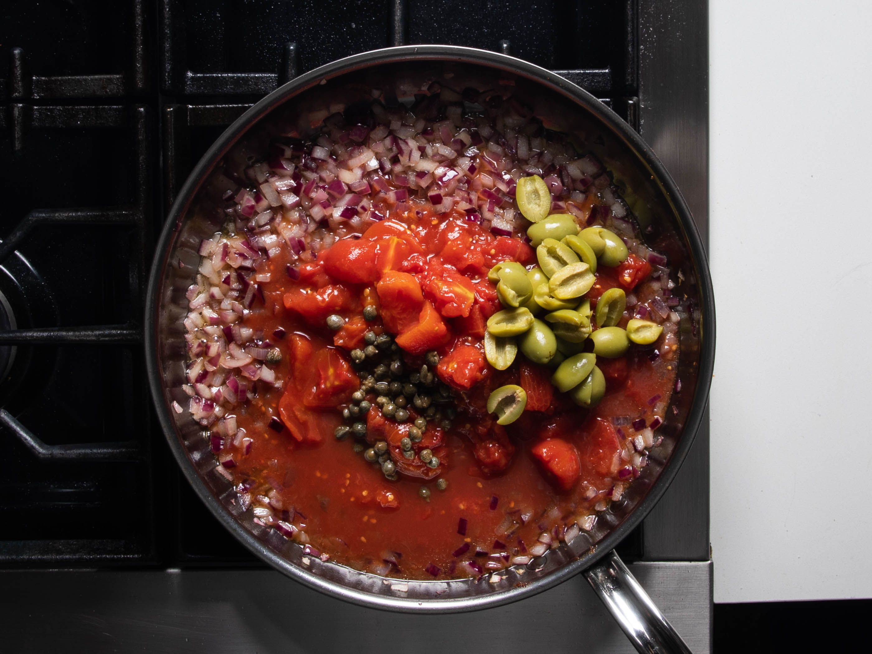 Tomato chunks and juices, olives, and capers have just been added to the pan on top of the sautéed onions, just before stirring together.