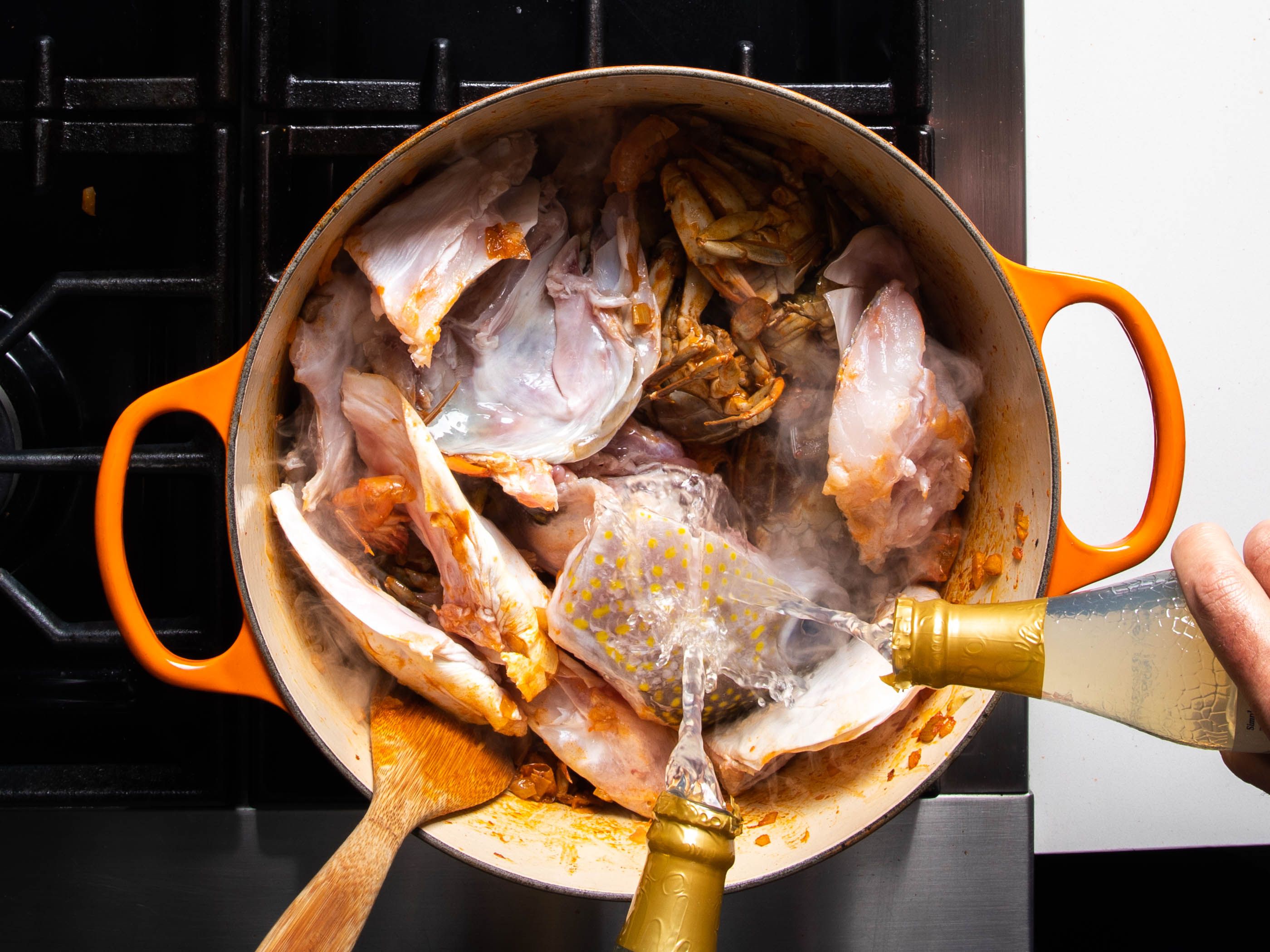 Clam juice is poured from the bottle into a pot that contains fish heads, crab bodies, tomato paste, and aromatic vegetables.
