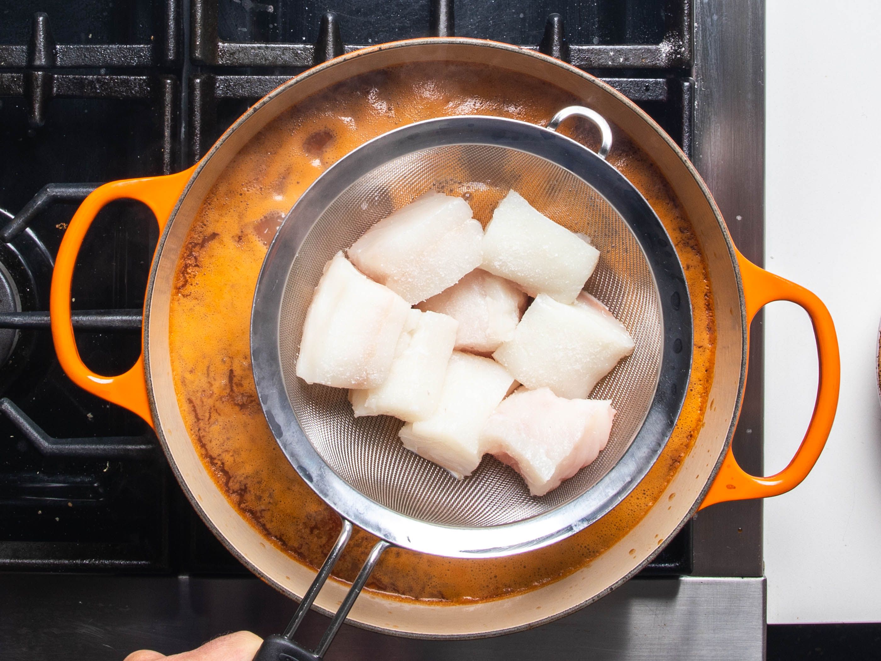 Portions of raw halibut sit in a fine mesh strainer suspended over a simmering pot of cioppino, ready to be dipped in and cooked.