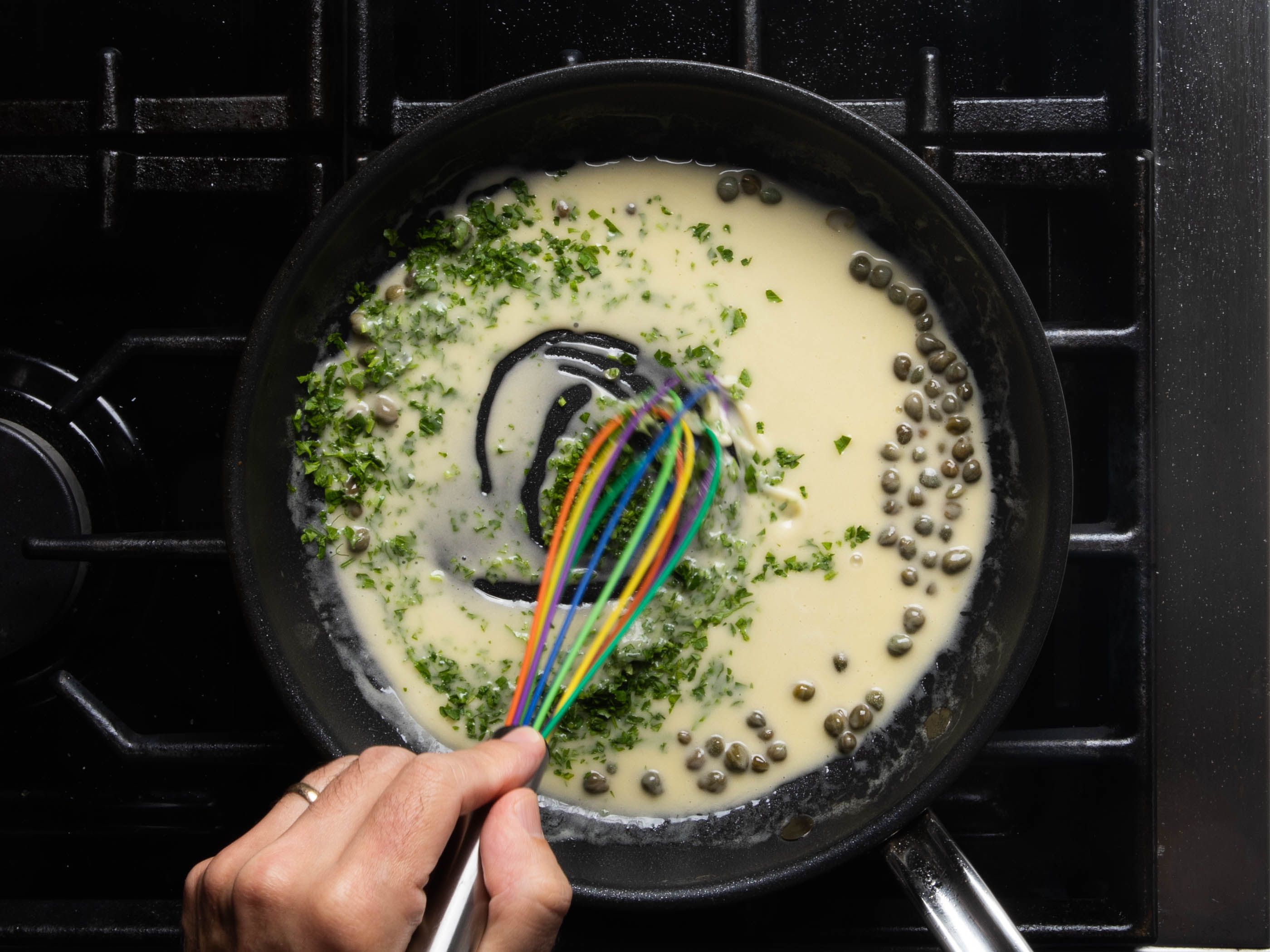 The pan sauce being whisked in the skillet with capers visible and the parsley just added; it's very creamy in appearance, and the whisk is leaving traces in the sauce, a sign of good viscosity.