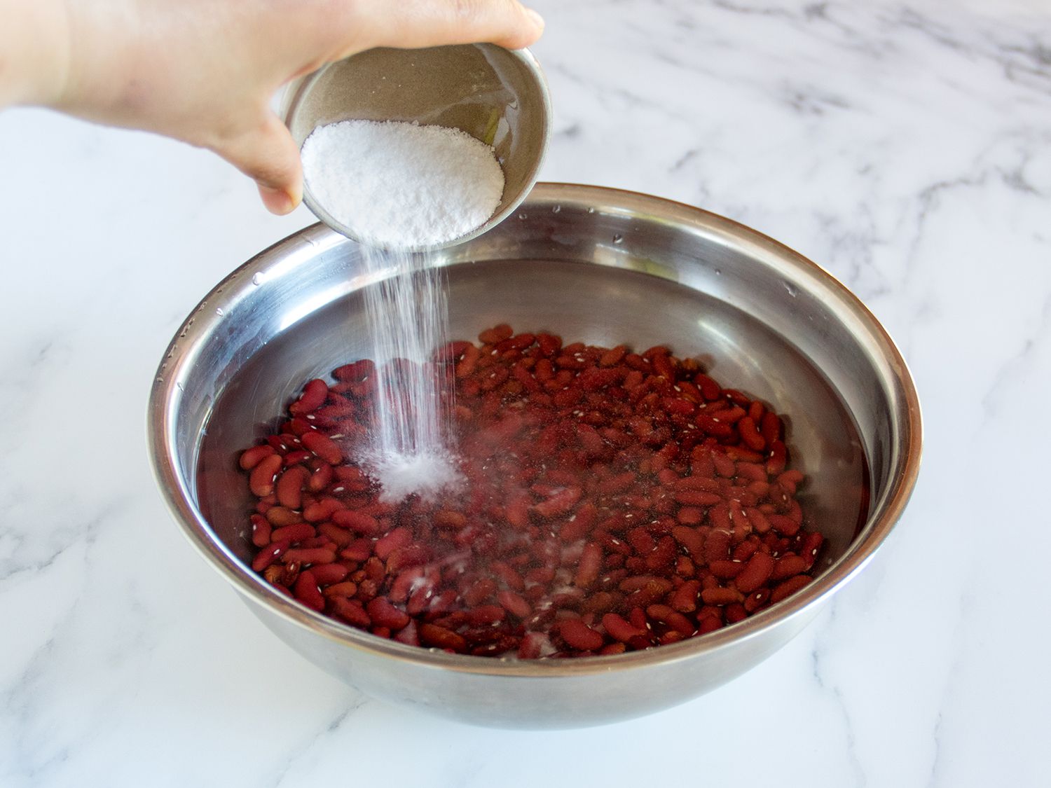Salt and baking soda being added to dried beans soaking in water in a mixing bowl