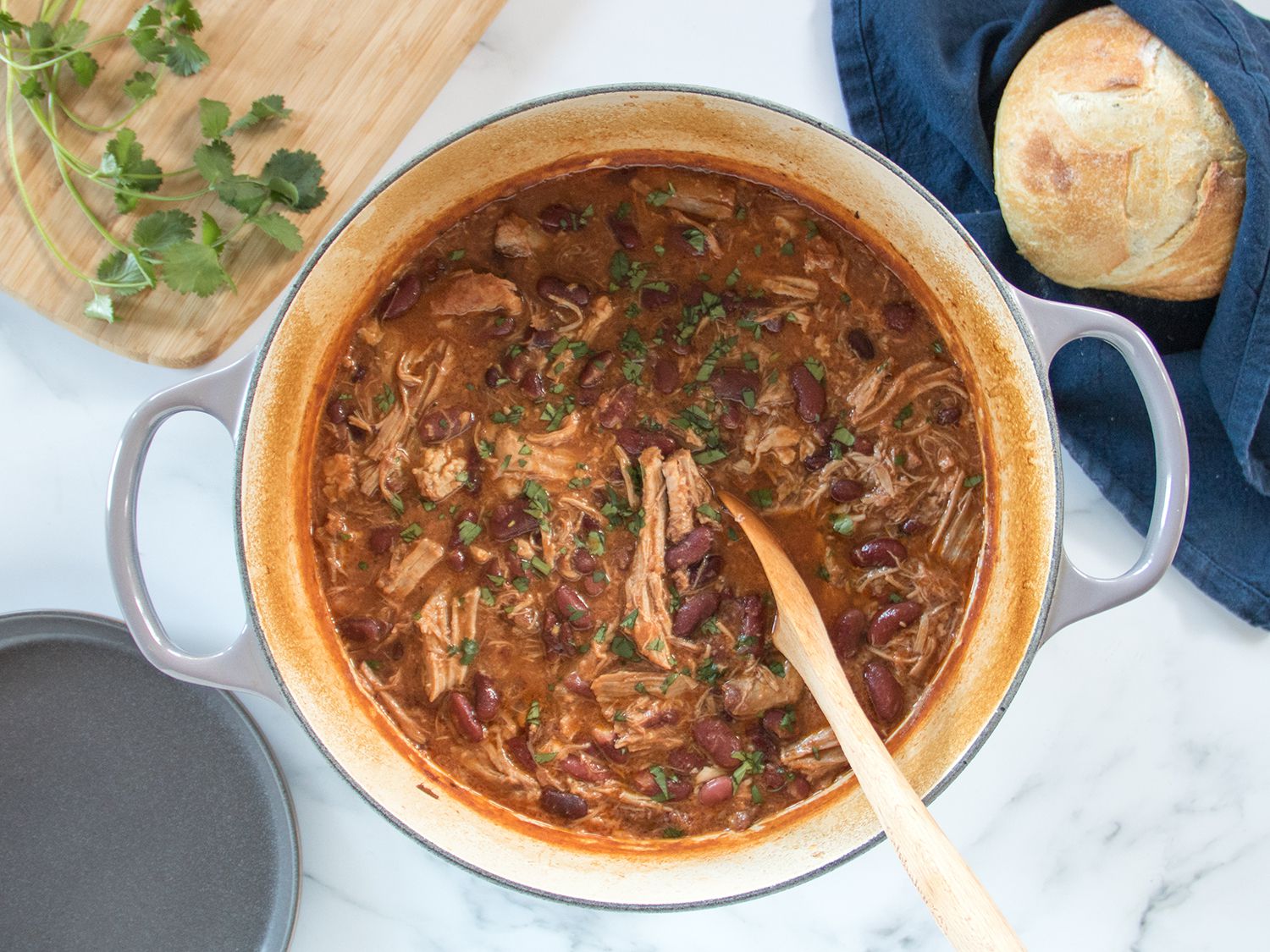 Pot of pork and creamy beans next to loaf of bread, a serving plate, and a cutting board