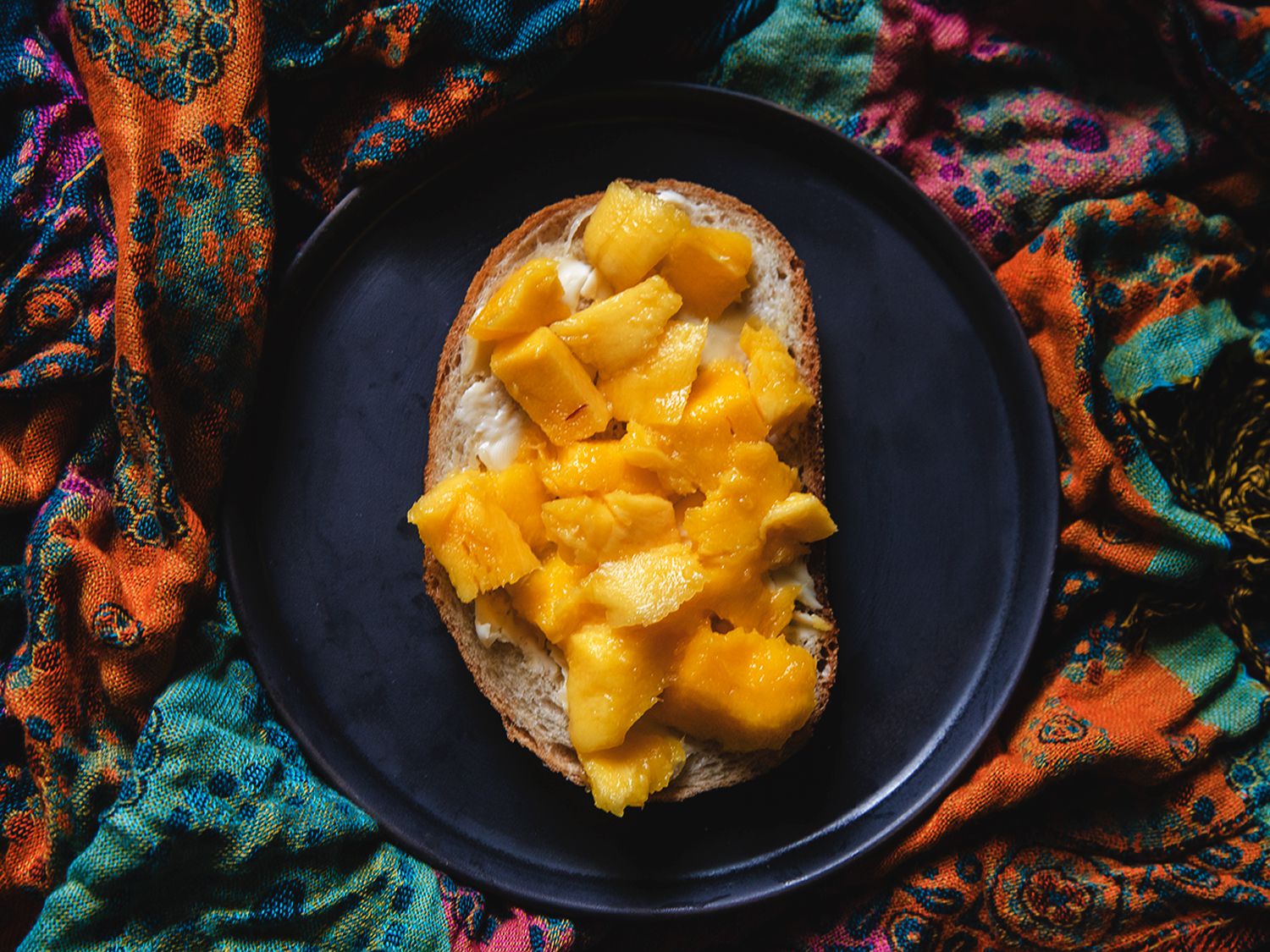 Mango on buttered toast on grey plate placed on top of a folded, brightly patterned background