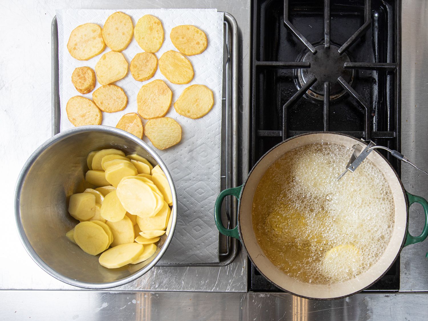 Overhead shot of potatoes being fried in a dutch oven with a bowl of sliced potatoes and potatoes resting on a paper towel.
