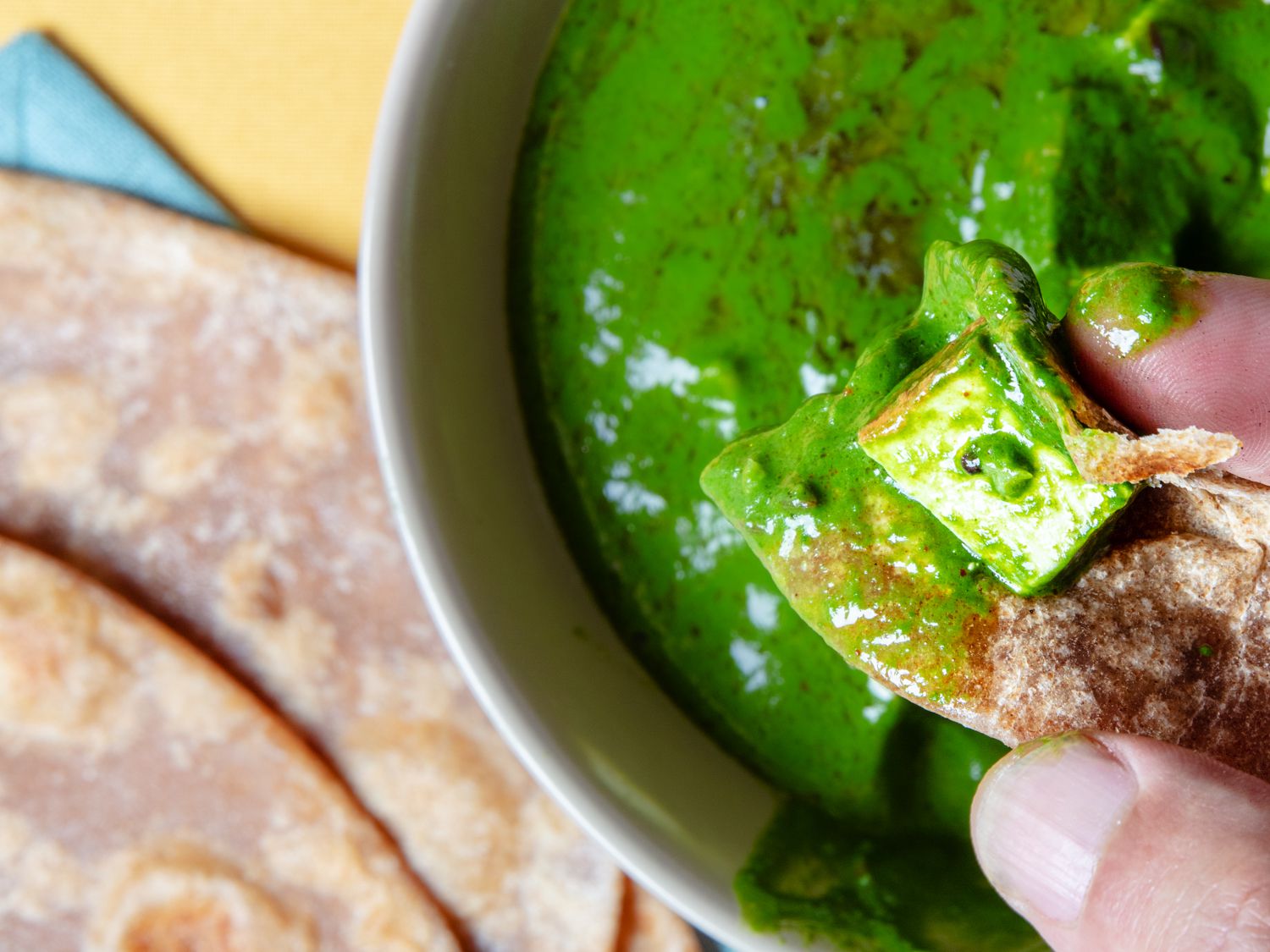 Close up of a hand holding a piece of roti with palak paneer on it