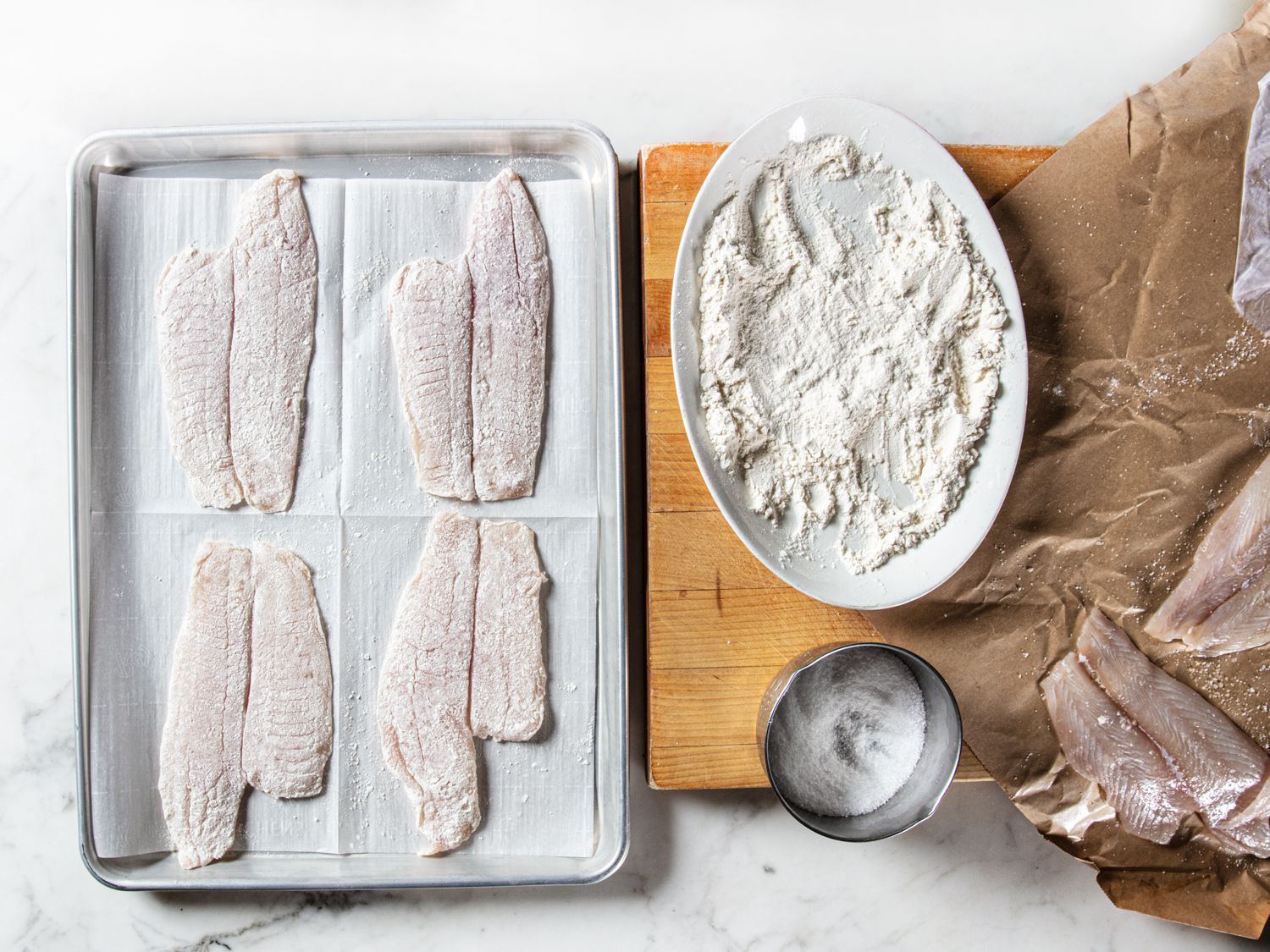 Four pieces of sole, dredged in flour on a baking sheet lined with parchment paper. Next to the baking sheet on a wooden cutting board is the plate with flour a bowl of salt and the remaining pieces of sole