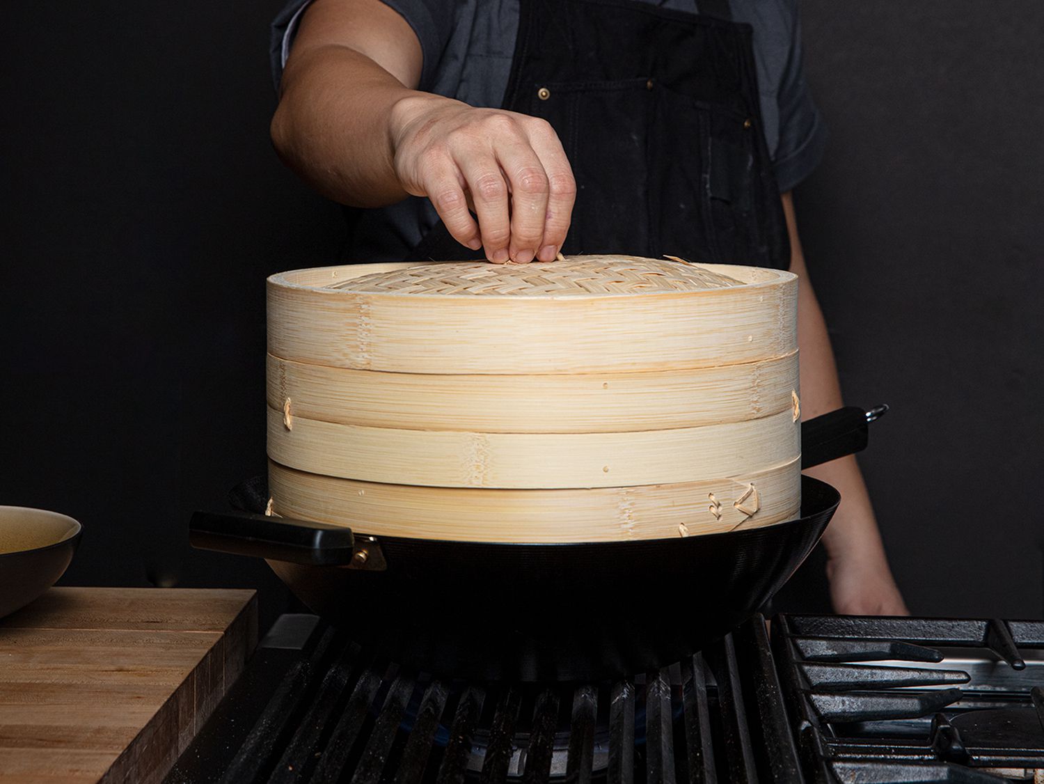 A person about to lift the lid off a four-tier bamboo steamer in a wok