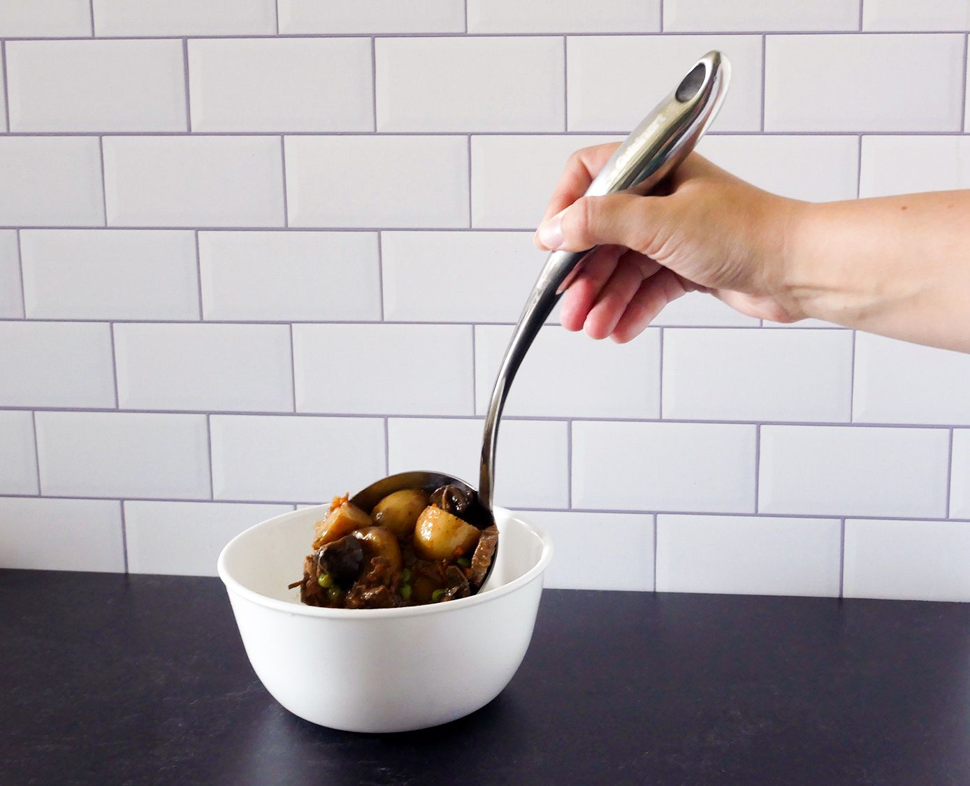 Hand using cuisinart ladle to pour stew into a white bowl.