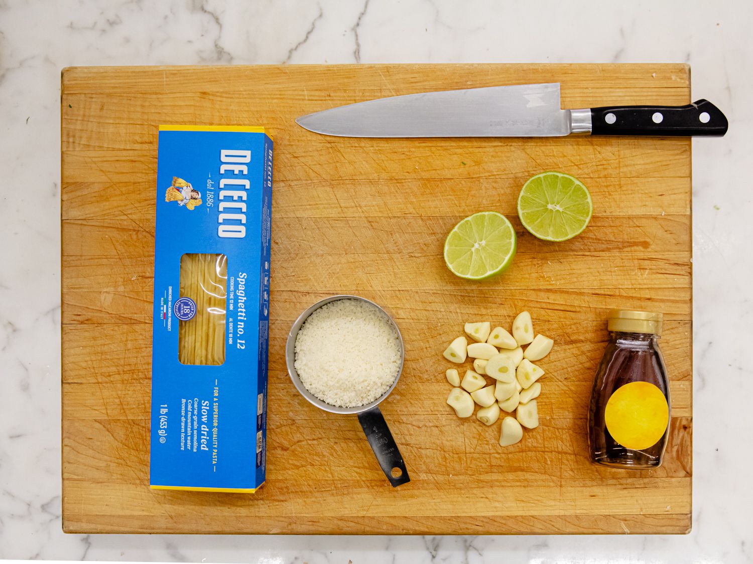 Overhead view of pasta, grated cheese, cut lime, garlic cloves, and honey on a cutting board
