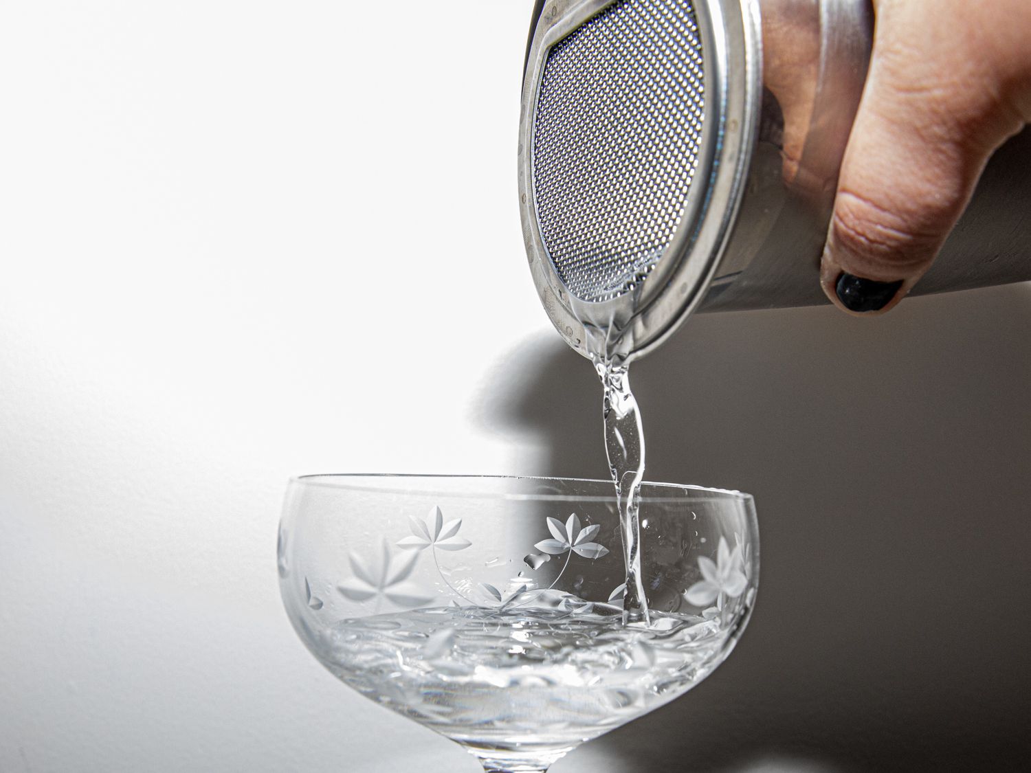 A hand pouring a cocktail into a coupe glass from a cocktail tin with an hawthorne strainer affixed to its opening
