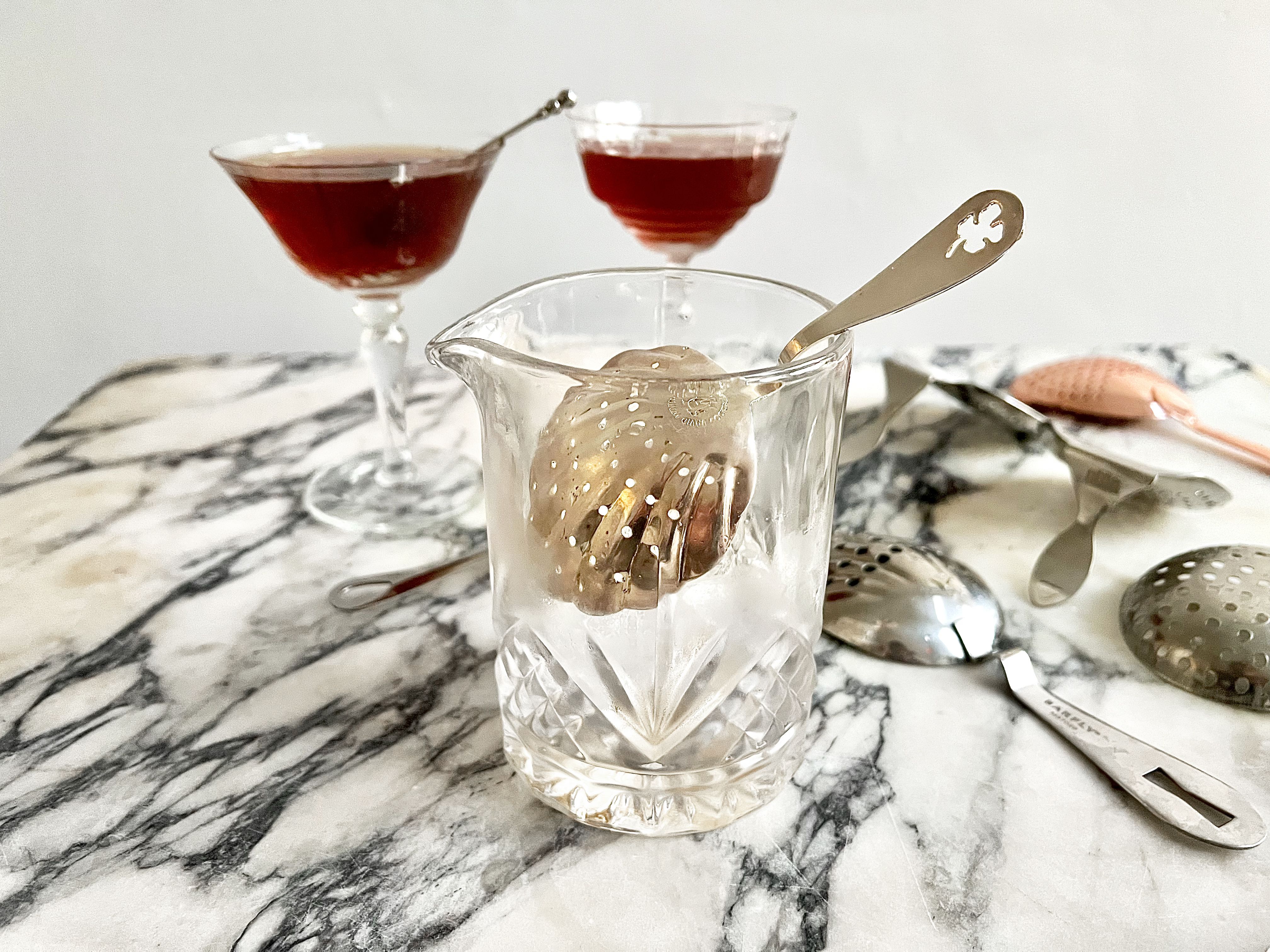 a julep strainer in a mixing glass with ice. behind it are two strained manhattans in coupe glasses.