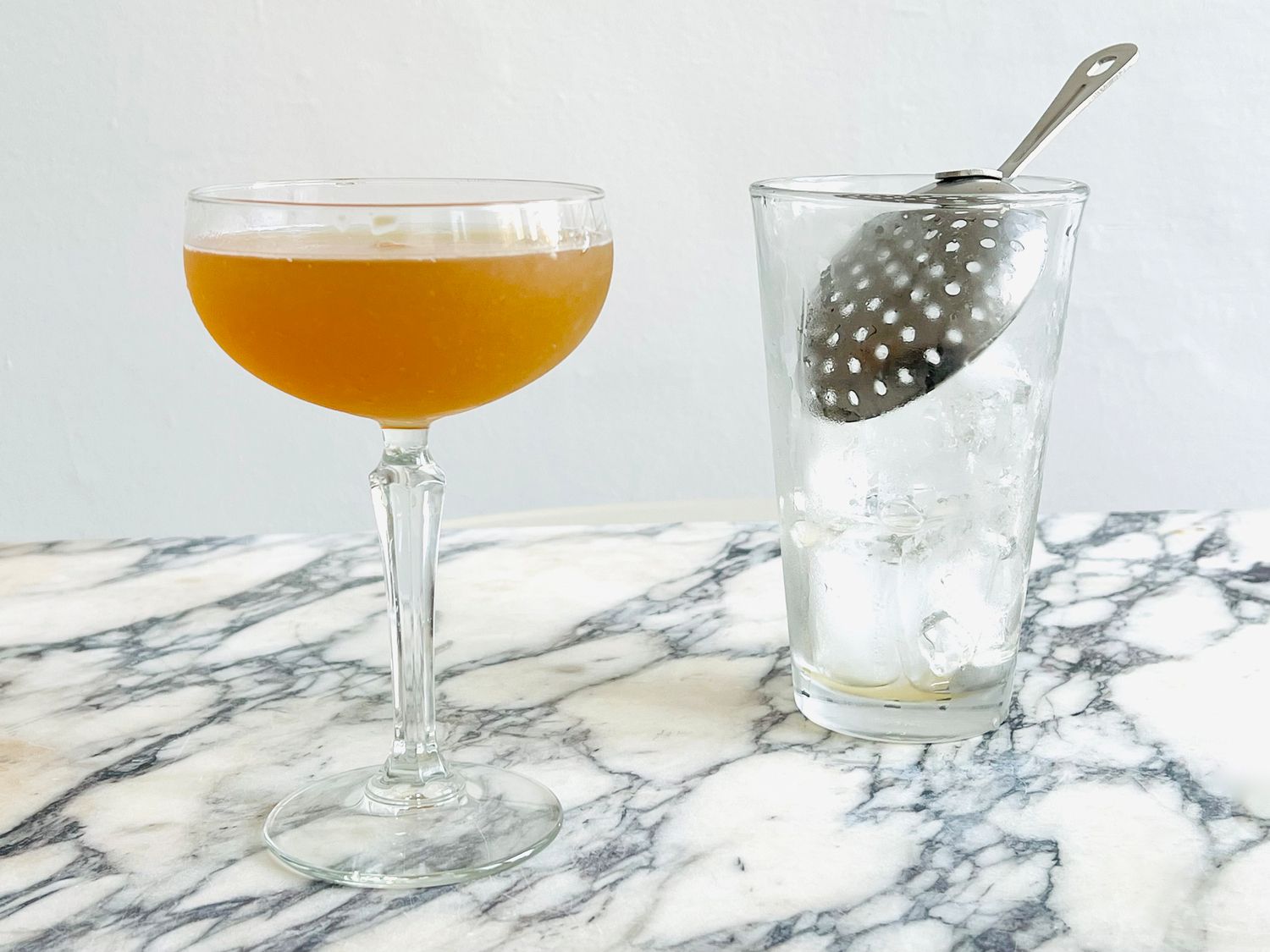 a straight handled julep strainer in a glass Boston shaker bottom with a cocktail in a coupe glass to the side.