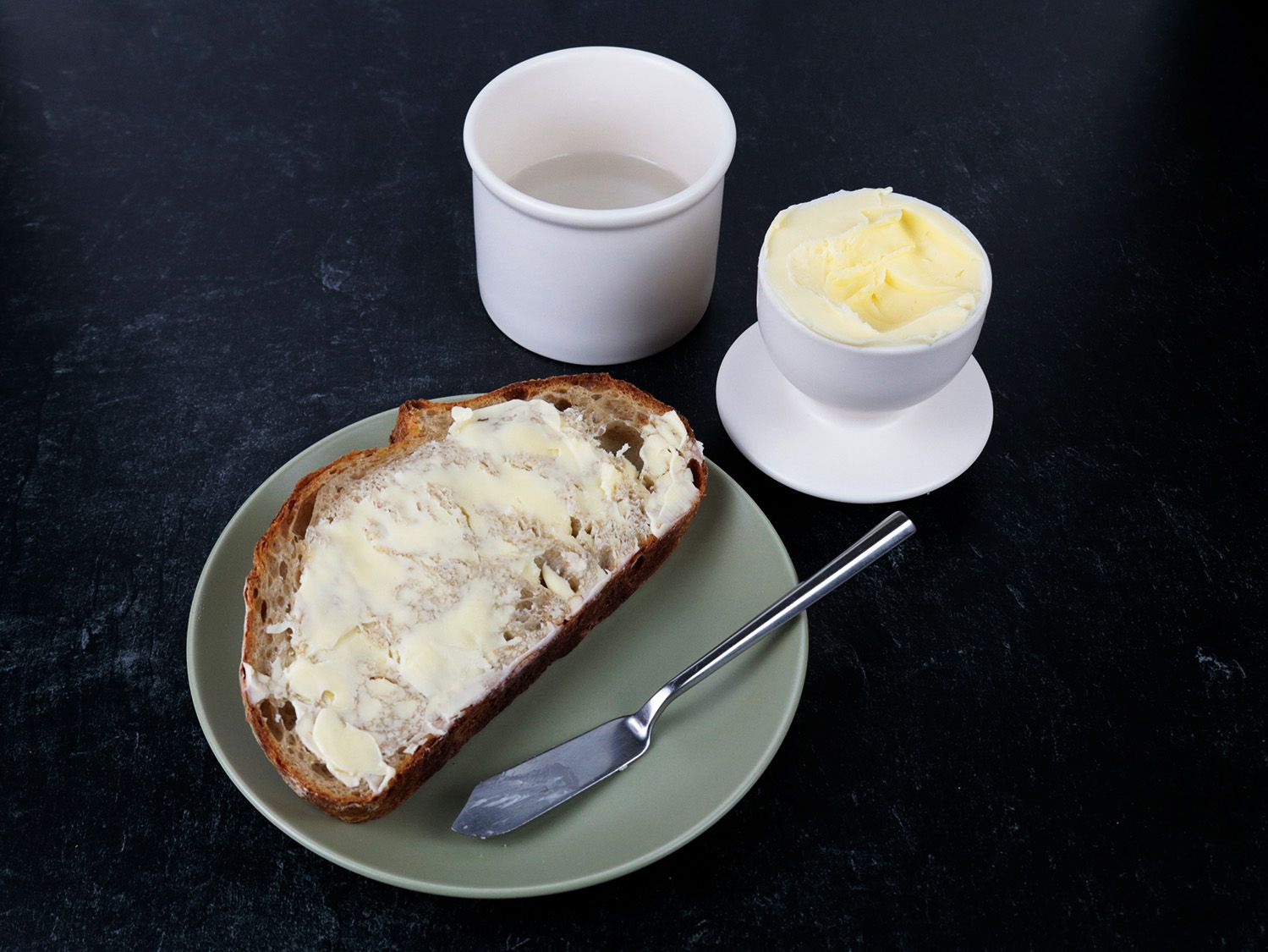 a slice of bread has a thick layer of butter on it resting on a plate near a butter bell ceramic crock