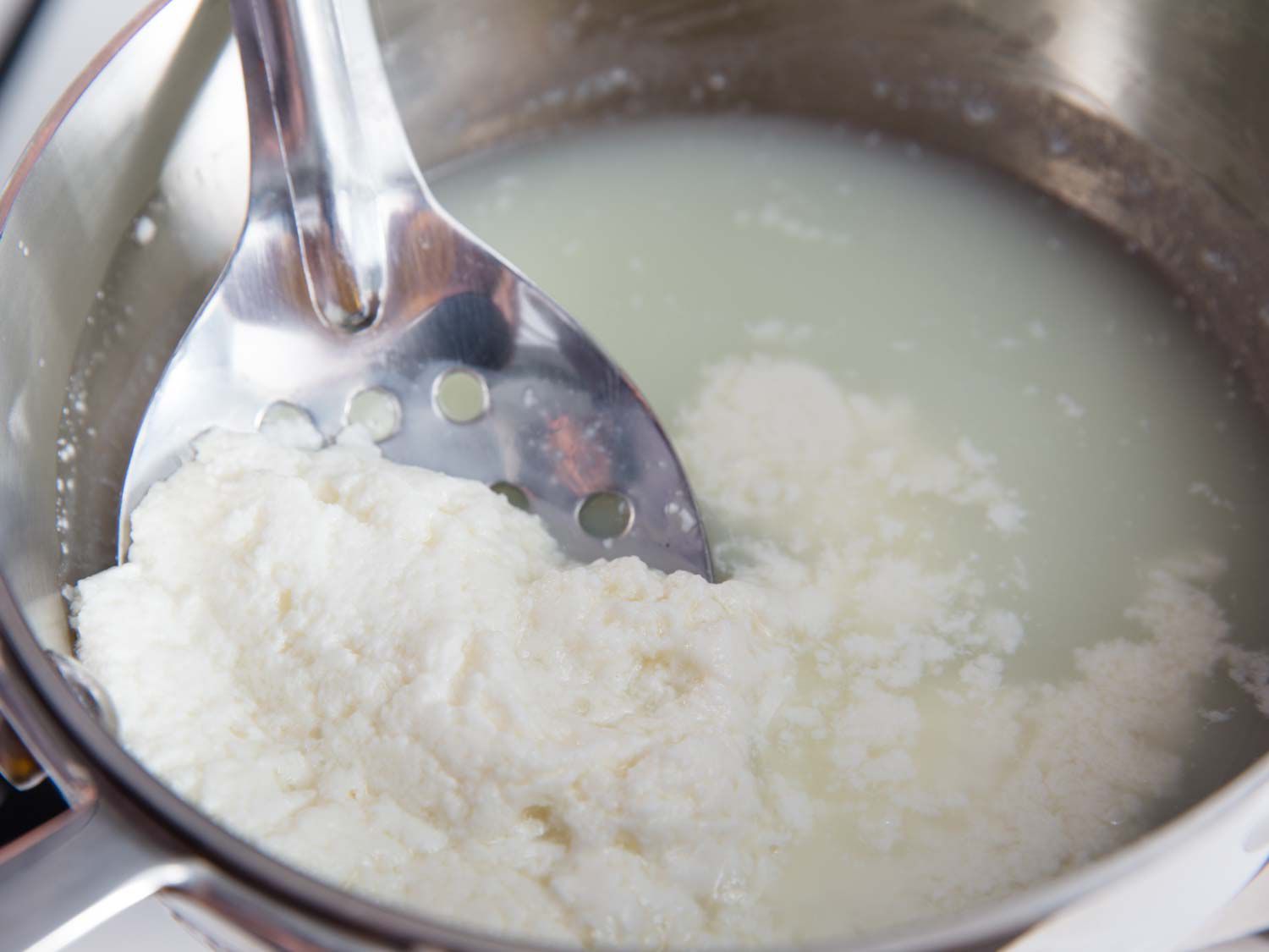 a slotted spoon removing homemade cheese from a saucepan