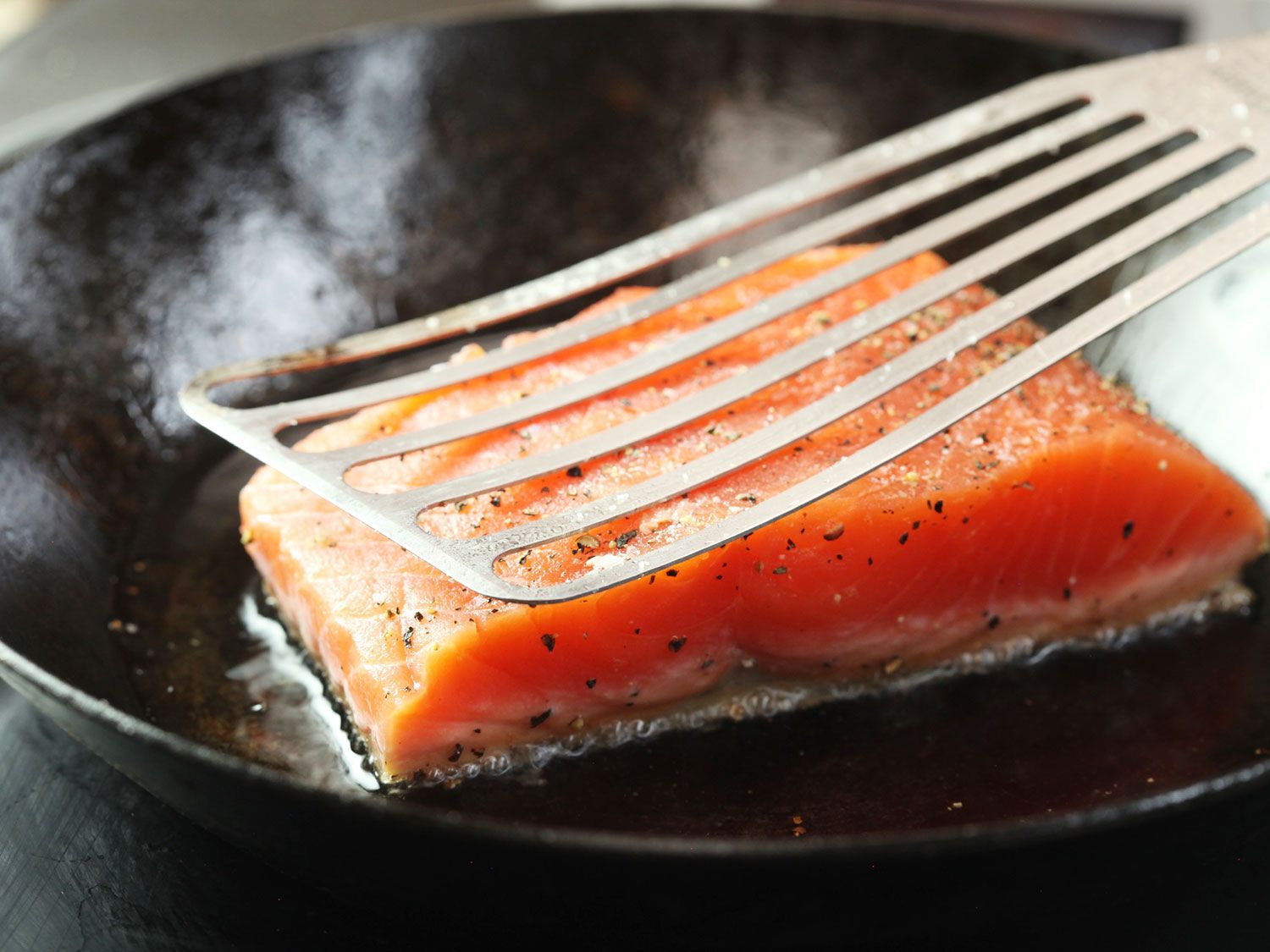 a fish spatula pressing down on a piece of salmon cooking in a carbon steel skillet