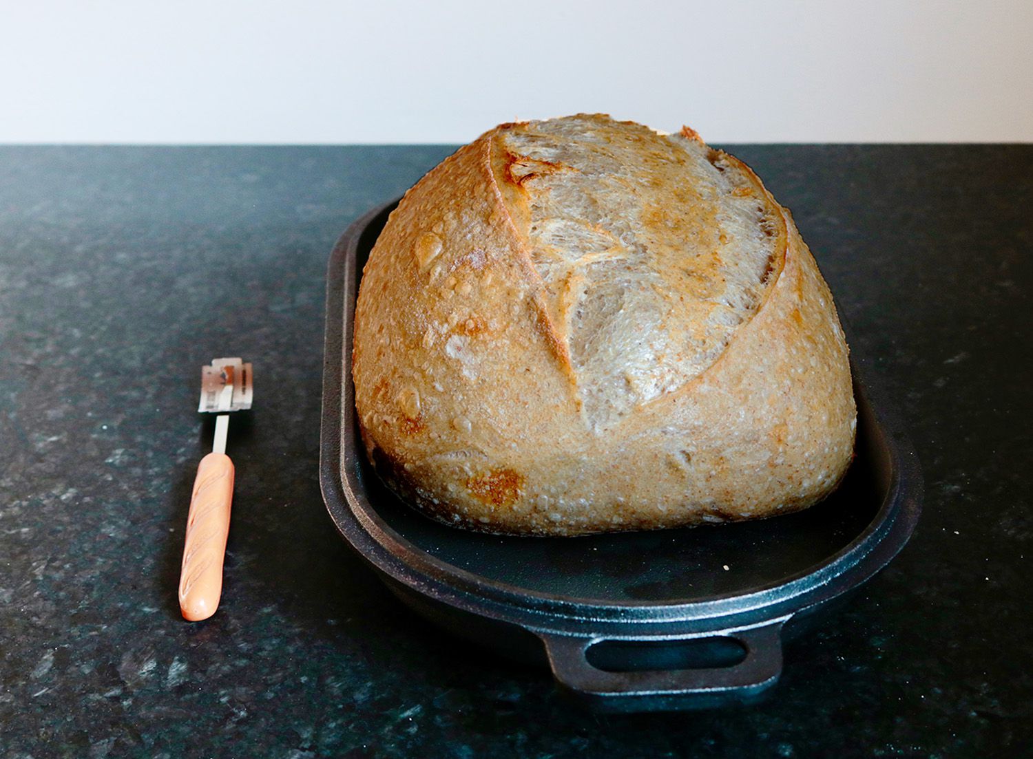 a partially baked loaf of bread rests on a cast iron pan next to a bread lame