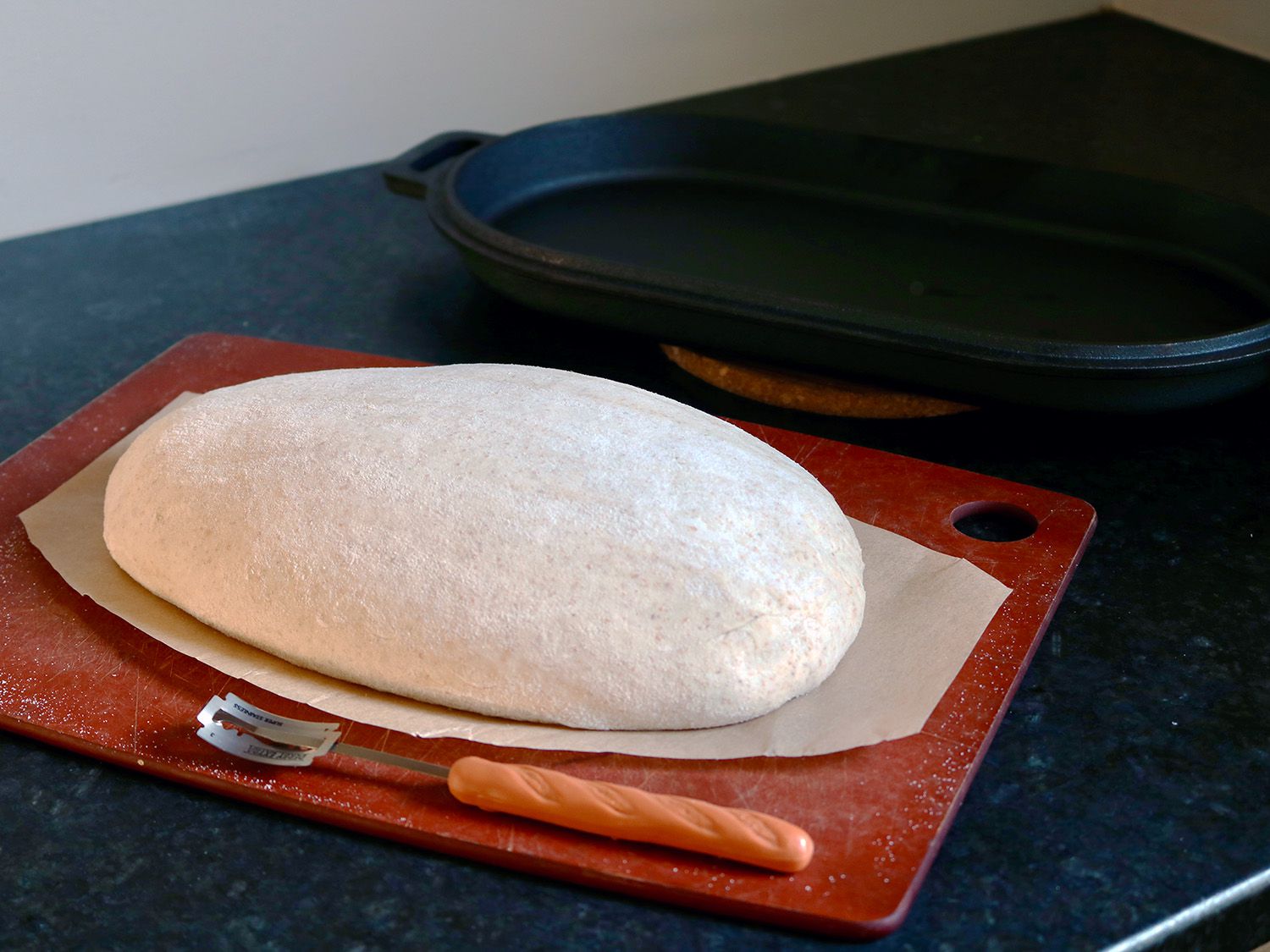 A loaf of bread dough rests on a cutting board next to a bread lame and the bottom of a cast iron pan.