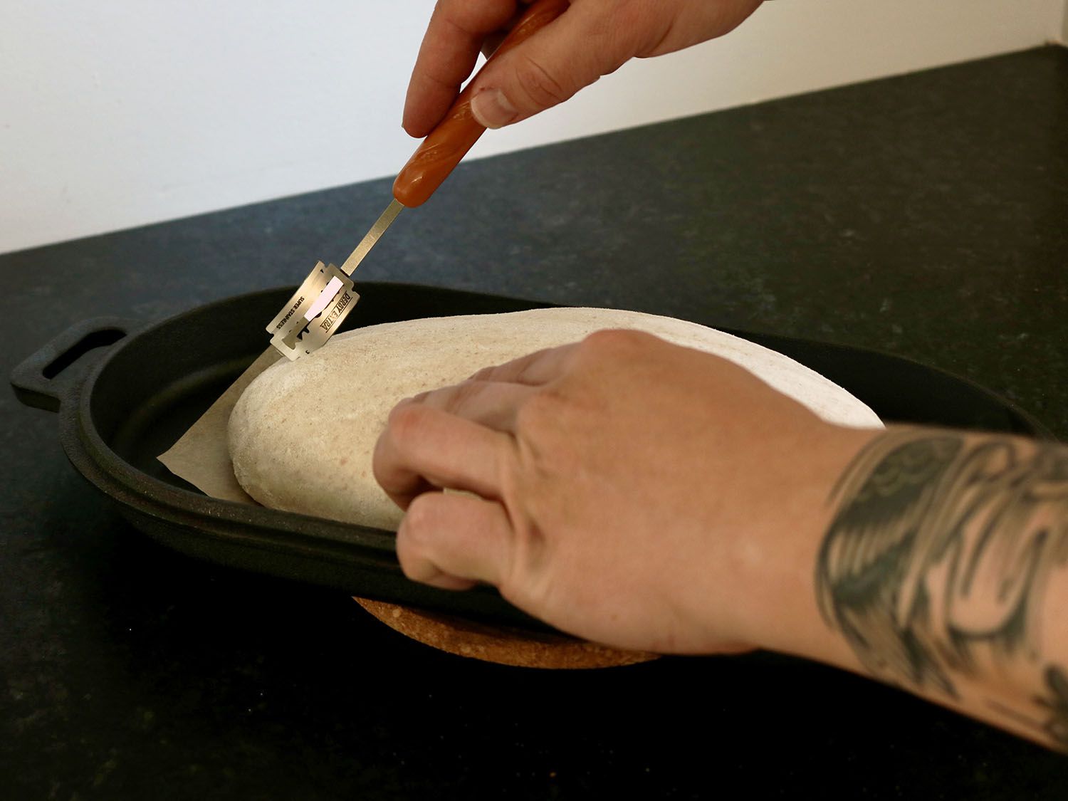 A bread dough is being slashed with a razor blade attached to a bread lame.
