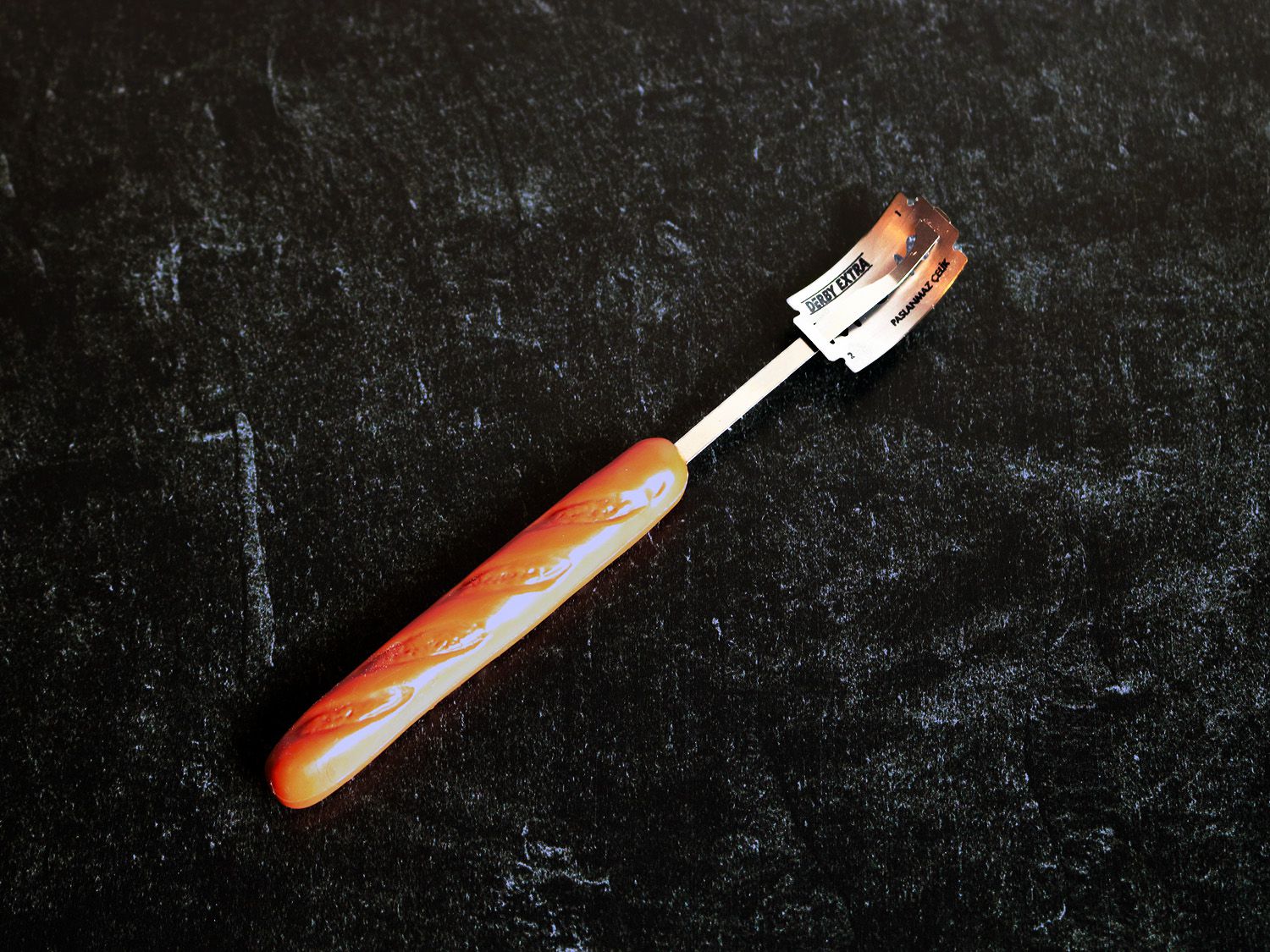 A baguette shaped handle attached to thin metal rod that holds a razor blade for scoring bread dough.