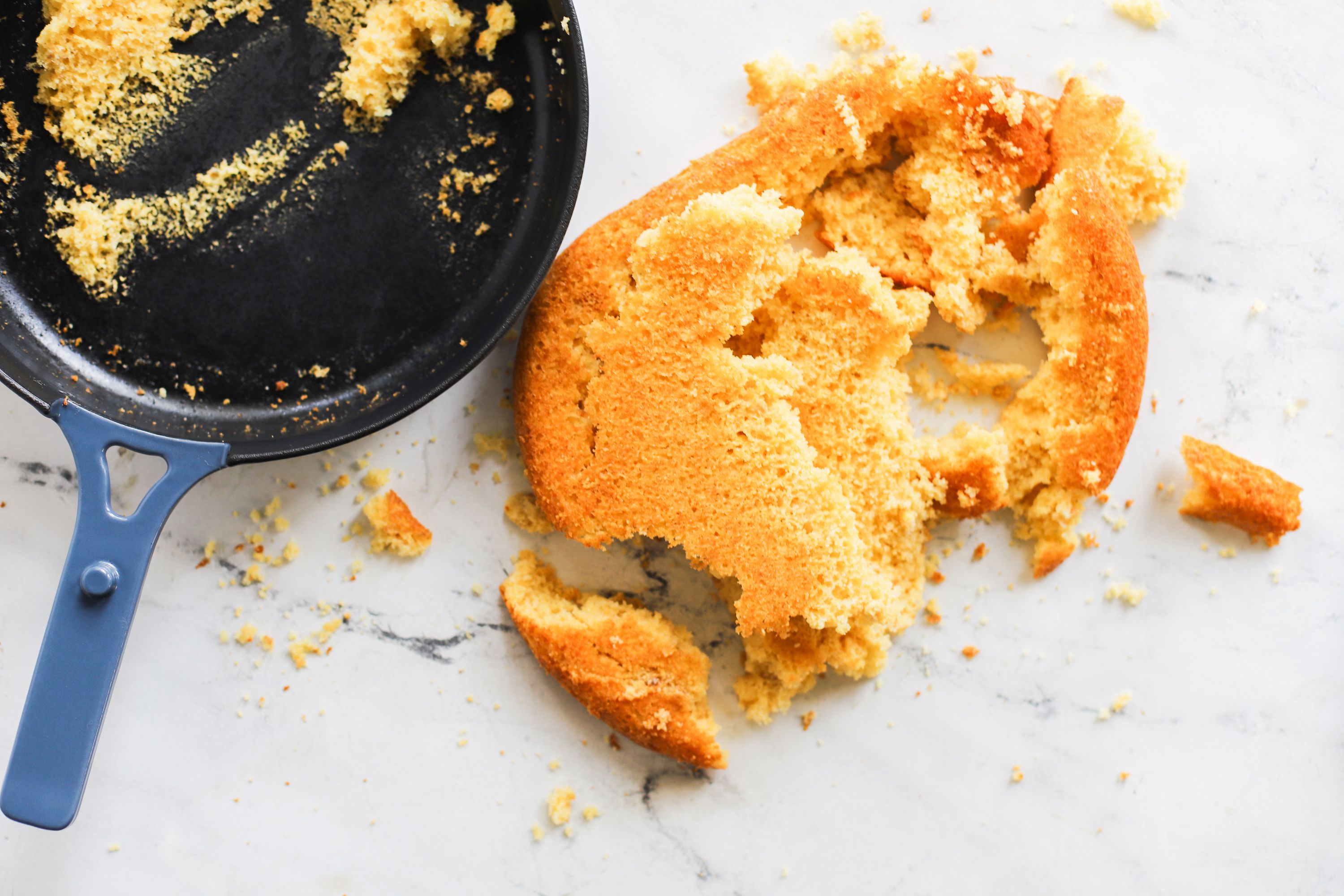 broken cornbread on a marble countertop with a skillet to the left of it.