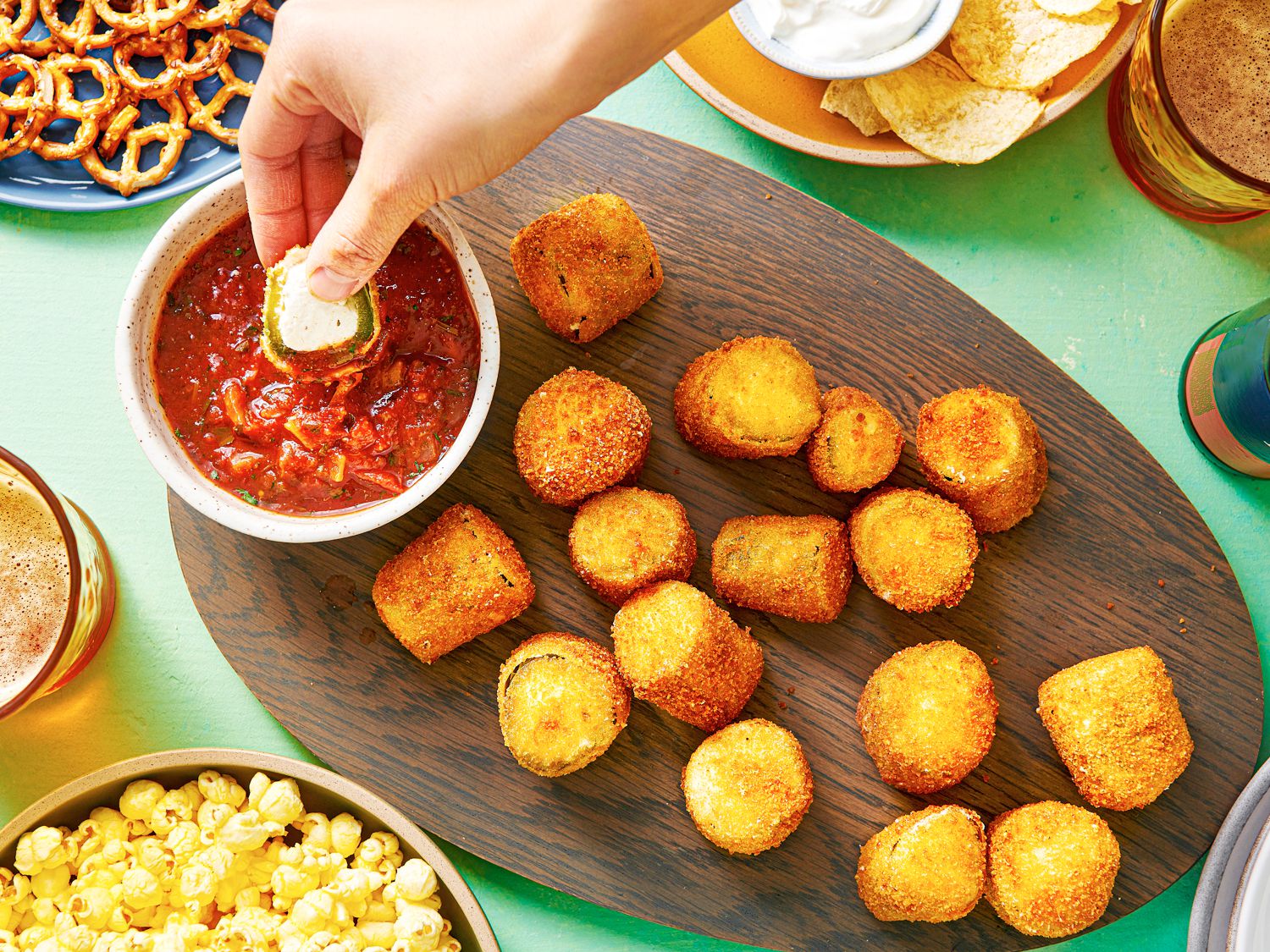 An oval wooden platter holding crispy deep-fried jalapeño poppers and a small bowl of tomato sauce. There is a hand dipping a popper into the tomato sauce, and the periphery of the image contains other snacks, such as pretzels, popcorn, and chips.