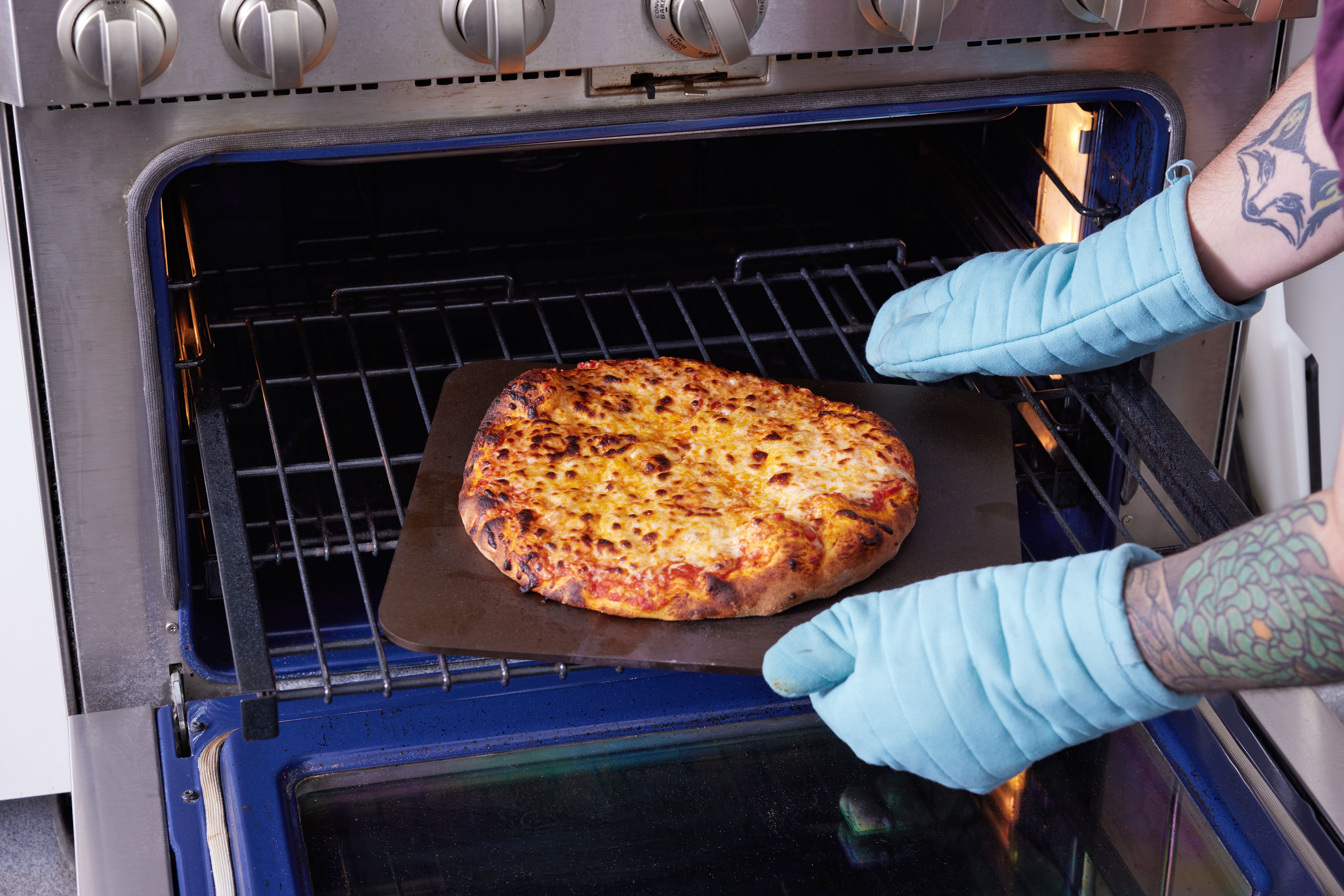 The Original Baking Steel being pulled out of the oven with a cooked pizza on it by a person wearing oven mitts 