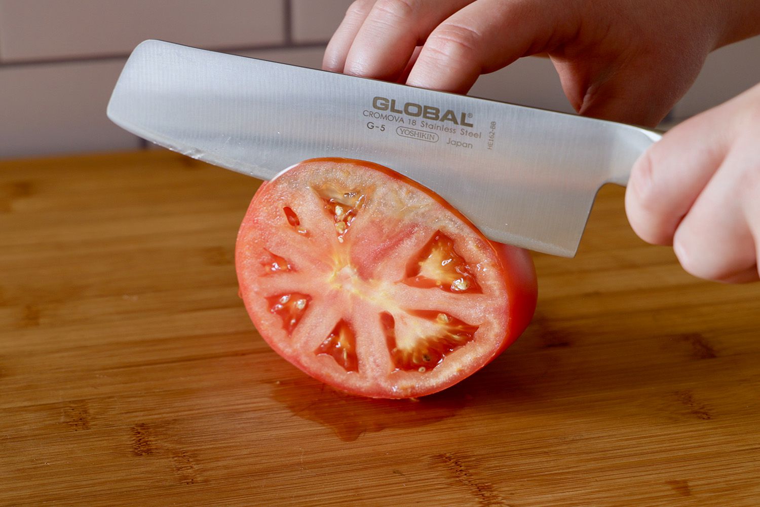 Two hands using a nakiri knife to thinly slice a tomato on a wooden cutting board