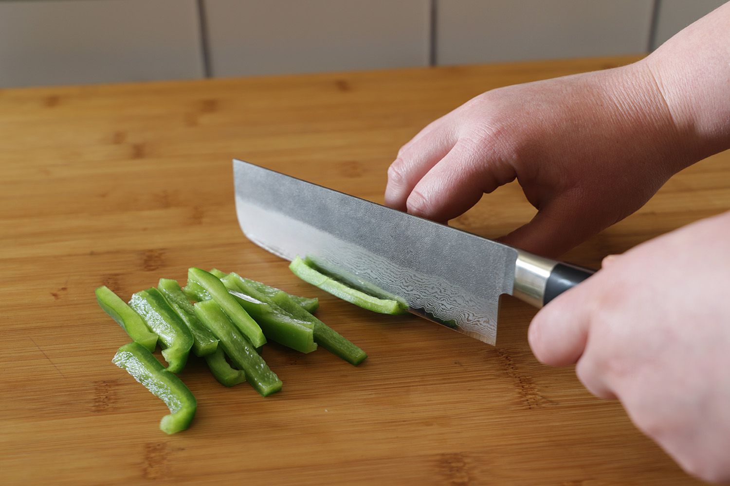 A person using a nakiri knife to thinly slice green bell pepper on a wooden cutting board