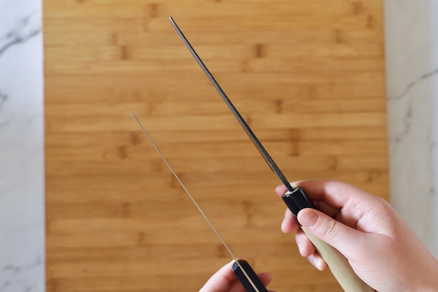 One hand holding a nakiri knife with a thicker blade and another hand holding a nakiri knife with a thinner blade