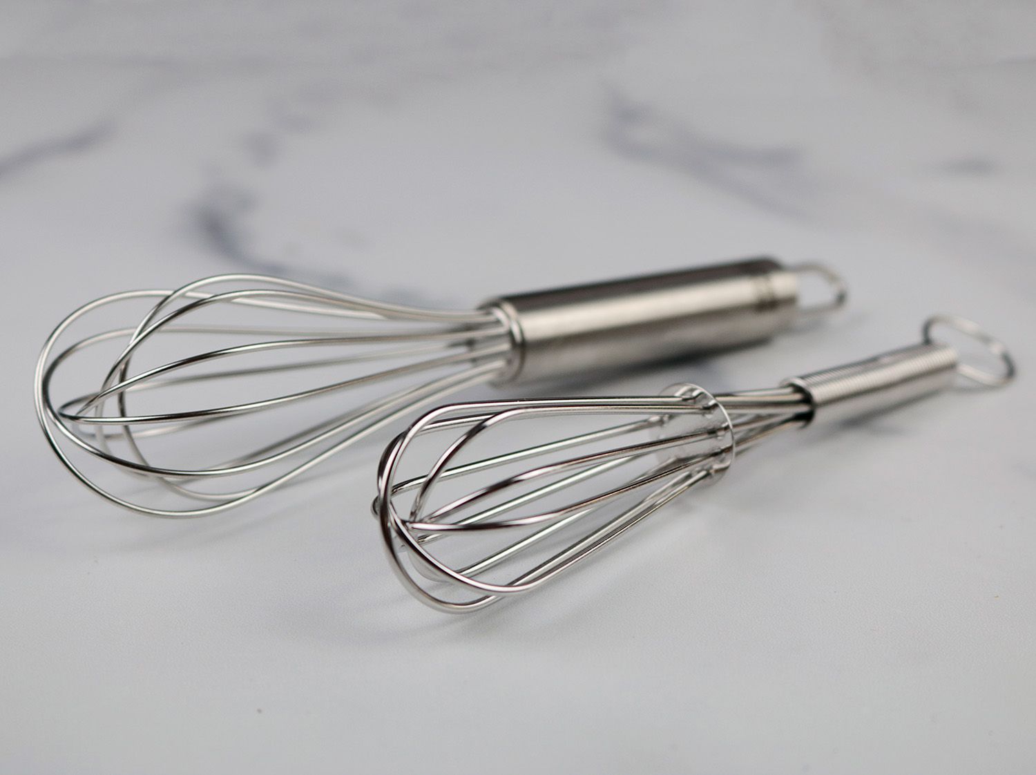 two miniature whisks, one longer than the other, on a white marble surface