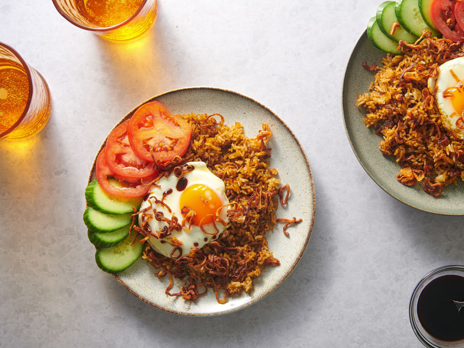 A plate of rice topped with a fried egg on a plate, garnished with cucumber and tomato slices, and showered with fried shallots. There is another plate to the right periphery of the image, as well as several glasses of liquid in the shot.
