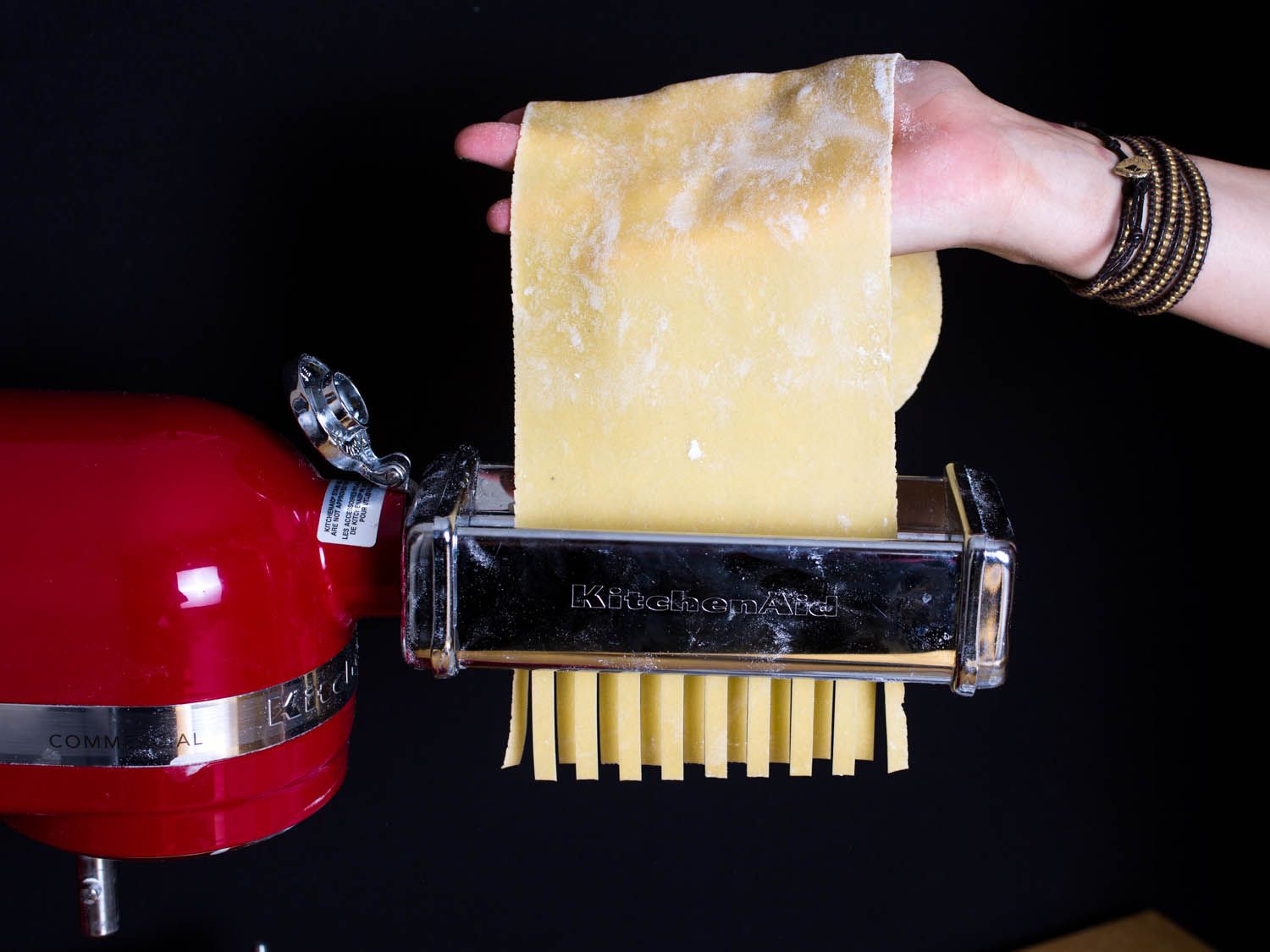 A hand feeding a sheet of pasta dough through the cutting attachment of a KitchenAid pasta maker