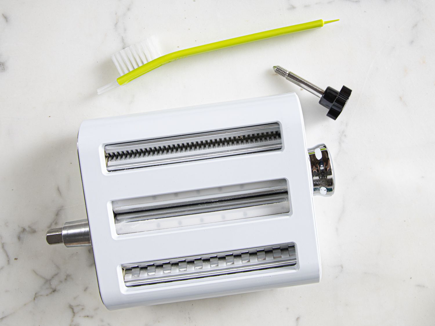 A white pasta roller and cutter stand mixer attachment on a marble surface with its cleaning brush beside it