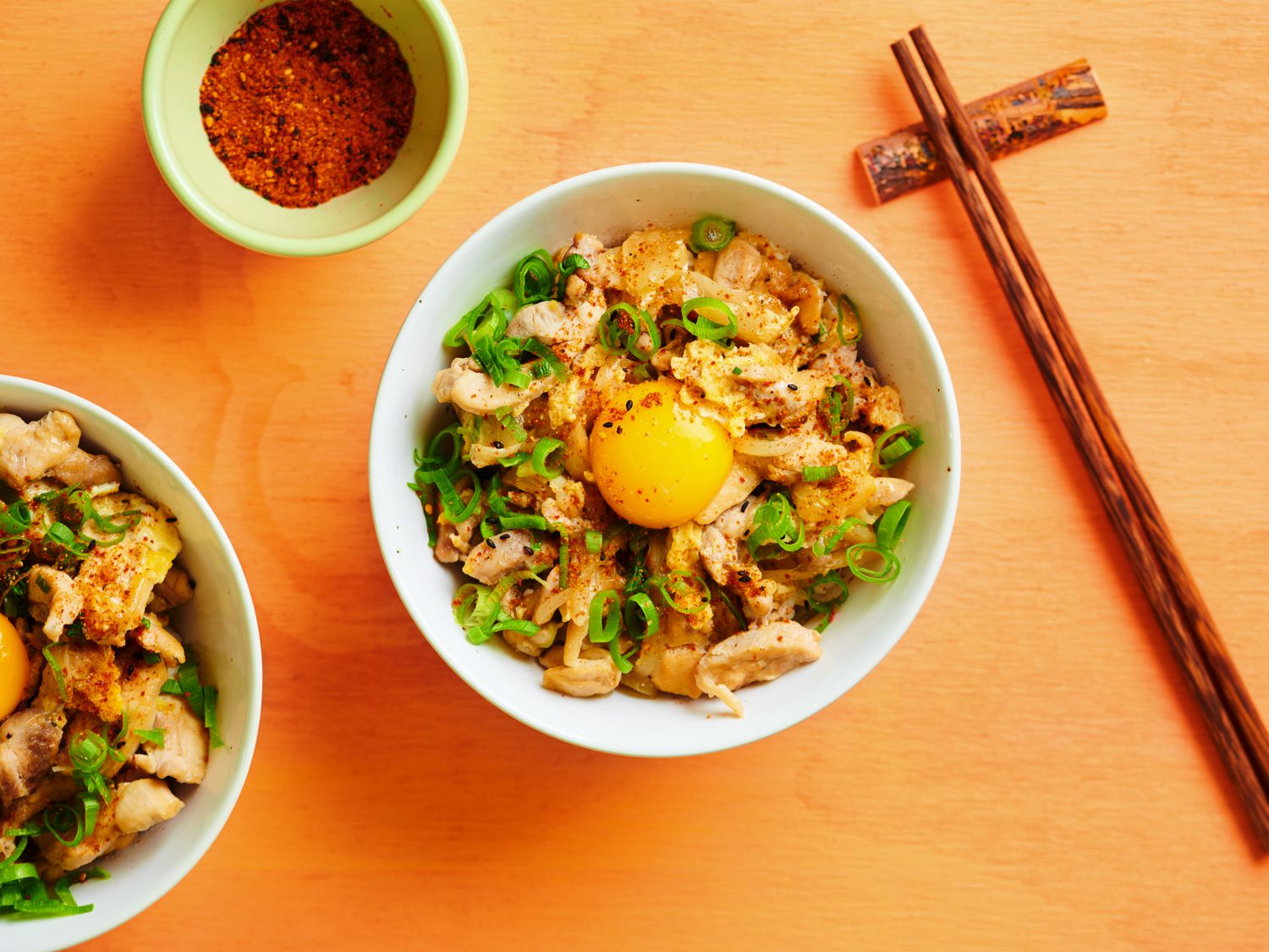 Oyakodon, in a white ceramic bowl alongside wooden chopsticks, a small ceramic bowl holding togarashi seasoning, and an additional bowl of oyakodon off to the left side.