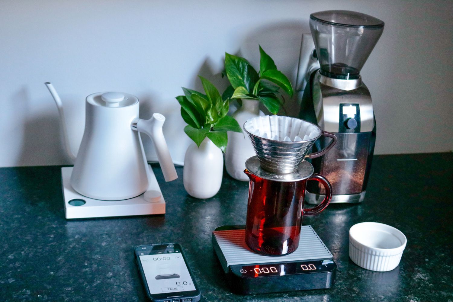 the acacia pearl kitchen scale on a counter with a coffee grinder, pourover maker, plant and orange glass cup