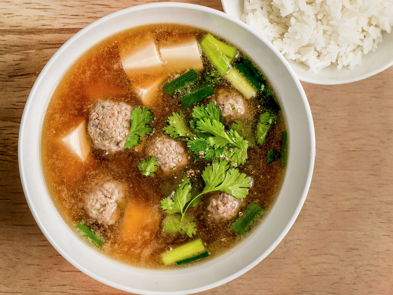 A white porcelain bowl holding the Thai pork and tofu soup. On the top righthand of the image is a bowl of white rice.