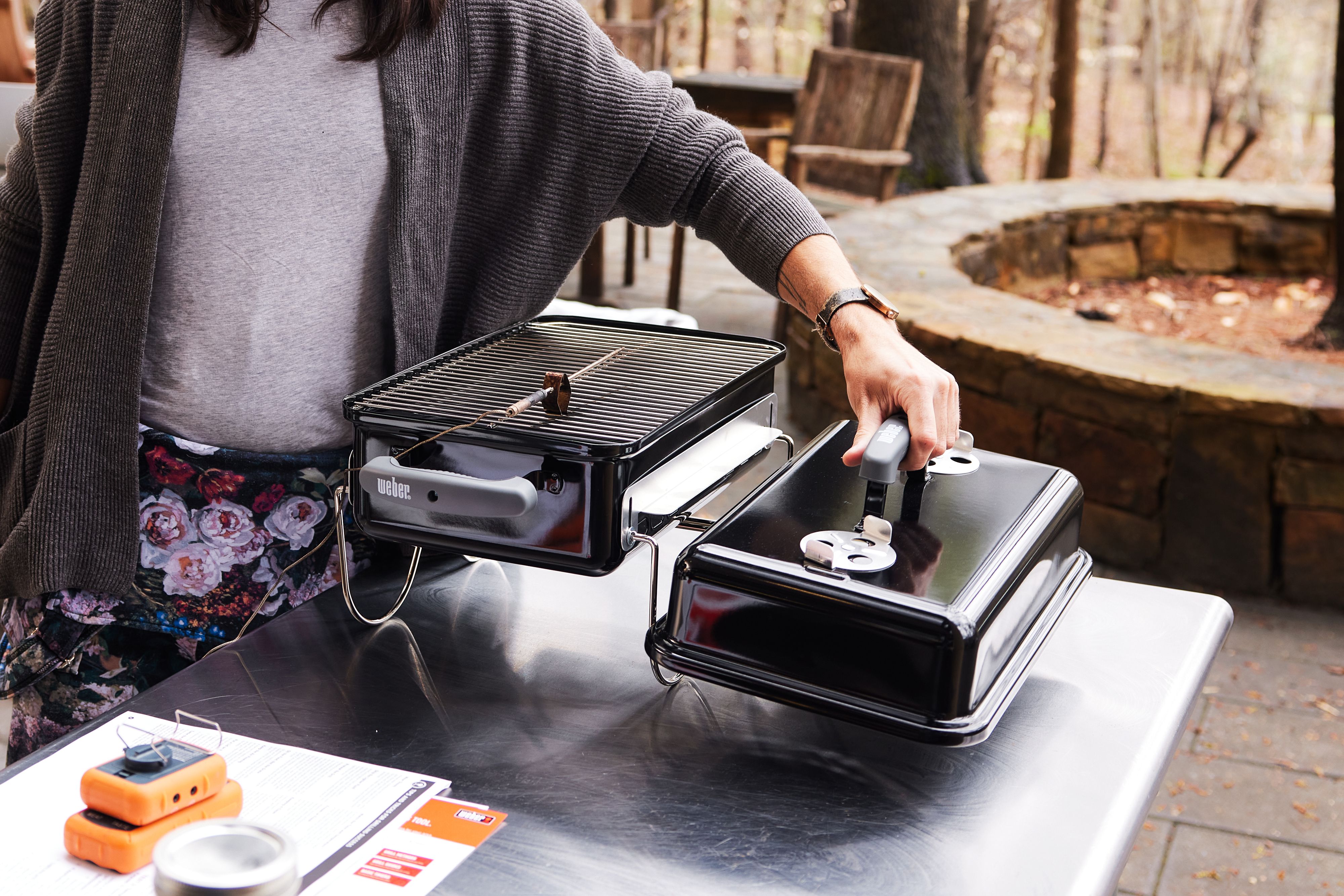 A person removing the lid of a portable charcoal grill that's set on a stainless steel table