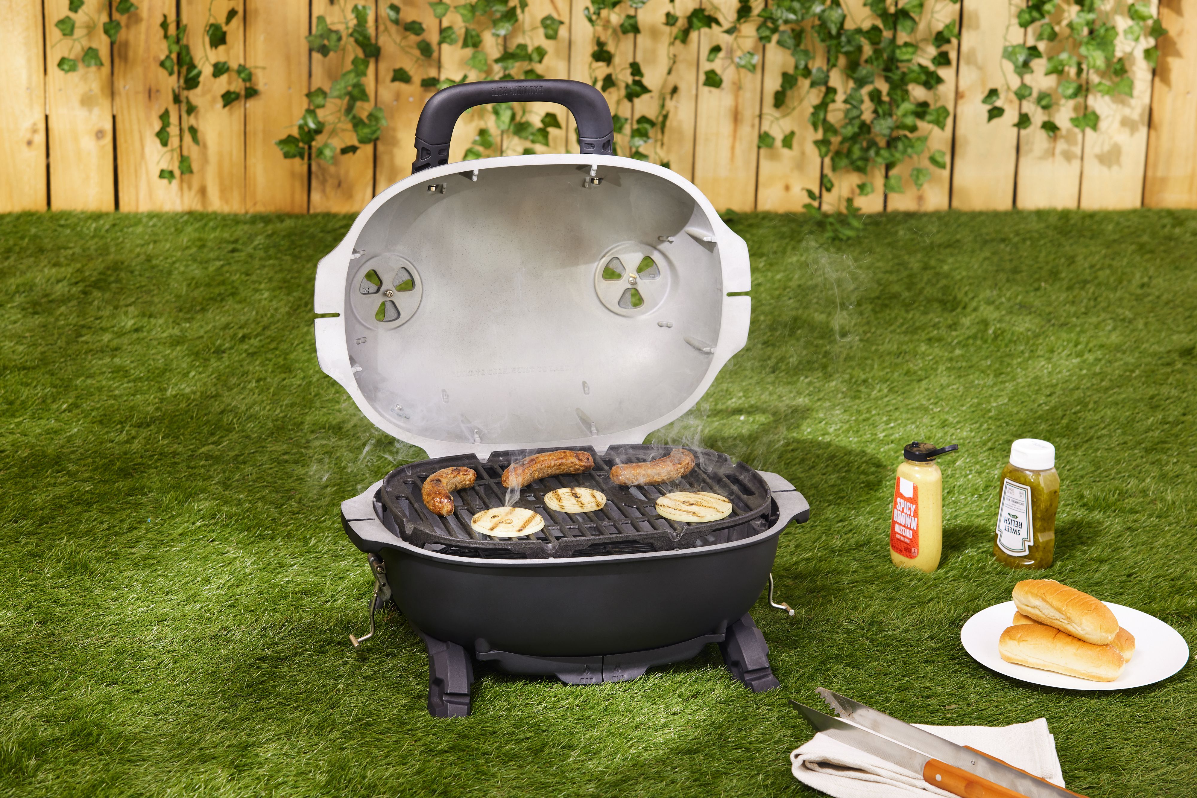 A portable charcoal grill cooking sausages, stationed on a lawn